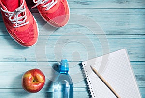 Fitness concept, pink sneakers, red apple, bottle of water and notebook with pencil on wooden background, top view