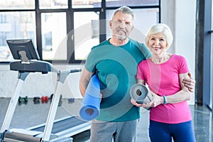 Fitness couple holding yoga mats and smiling at camera in gym