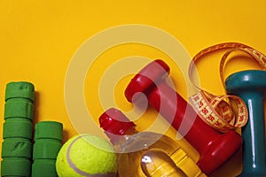 Fitness and diet concept background. Tennis ball with dumbbells and bottle of water isolated on yellow background