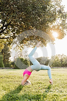 Fitness exercises on the grass. Stretching before a workout at sunset. Sports girl stands on her head near a tree in the