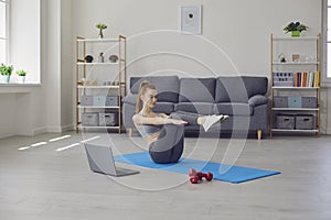 Fitness girl doing exercises on the floor and watching a training video while having a laptop in a living room. Online