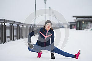 Fitness girl model runner doing flexibility exercise for legs before run at snow winter promenade