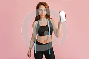 Fitness girl smiling and holding a phone with a mockup and posing hold measure tape in black sportswear on a pink background. Slim
