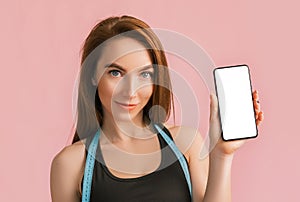 Fitness girl smiling and holding a phone with a mockup and posing hold measure tape in black sportswear on a pink background. Slim