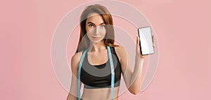 Fitness girl smiling and holding a phone with a mockup and posing hold measure tape in black sportswear on a pink background. Slim