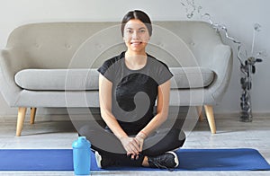 Fitness at home concept. Smiling young asian woman is sitting on mat with sports equipment at home