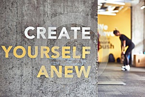 Fitness motivation quote Create Yourself Anew written on grey beton wall at gym, people working out on the background