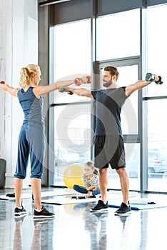 Fitness people exercising with dumbbells while teenage girl sitting on mat