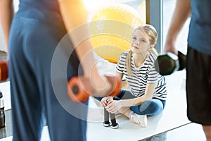 Fitness people exercising with dumbbells while teenage girl sitting on mat