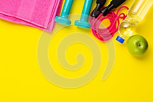 Fitness and sport concept. Jump rope, green apple, dumbbells, bottle of water and pink towel on bright yellow background. Free