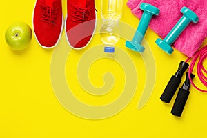 Fitness and sport concept. Jump rope, green apple, dumbbells, bottle of water, red sneakers and pink towel on bright yellow