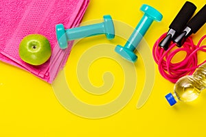 Fitness and sport concept. Skipping rope, green apple, dumbbells, bottle of water and pink towel on bright yellow background. Free