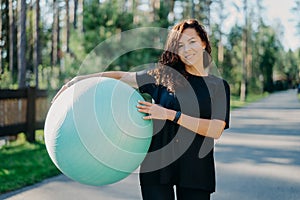 Fitness, sport and healthy lifestyle concept. Smiling curly brunette woman in black t shirt holds big fitness ball poses outside