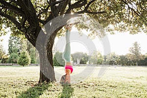 Fitness stretching exercises before workout. Sports girl stands on her head near a tree in the park.