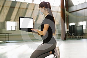 Fitness trainer kneel down in empty gym. Woman in medical mask works remotely indoor in solitude. She sits with laptop