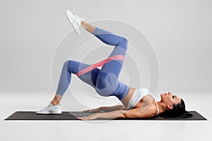 Fitness woman doing glute bridge exercise with resistance band on gray background. Athletic girl working out