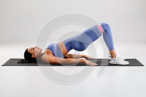 Fitness woman doing glute bridge exercise with resistance band on gray background. Athletic girl working out