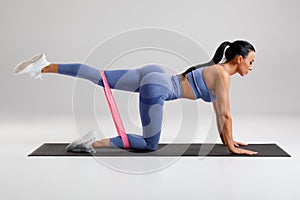 Fitness woman doing kickback exercise for glutes with resistance band on gray background. Athletic girl working out donkey kicks