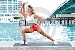Fitness woman doing lunges exercises for leg muscle and shoulders training with resistance band outdoors