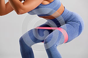 Fitness woman doing squats exercise for glute with resistance band on gray background. Athletic girl working out