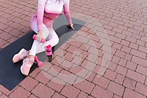 FItness woman in pink sportswear sits on yoga mat with dumbbell in hands.