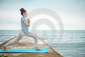 Fitness woman practicing yoga virabhadrasana posture hands namaste on pier dock sea landscape