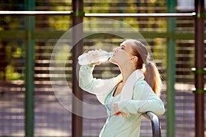 Fitness woman resting after workout, relaxing, drinking clear water from bottle outdoor fitness area in a park