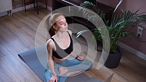 Fitness woman sitting on yoga mat, doing stretching exercise following online course on laptop.