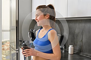 Fitness woman standing with black water bottle in kitchen, wearing workout clothing, drinking after yoga exercises