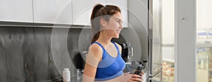 Fitness woman standing with black water bottle in kitchen, wearing workout clothing, drinking after yoga exercises