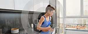Fitness woman standing with black water bottle in kitchen, wearing workout clothing, drinking after yoga exercises