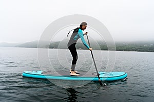 Fitness woman standing on surfboard enjoying supsurfing with paddle at endless sea water fog