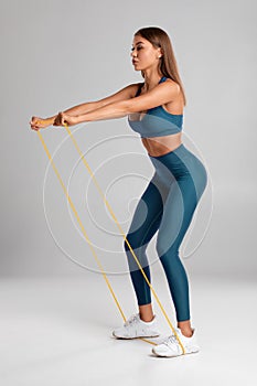 Fitness woman using a resistance band in her exercise routine. Sexy athletic girl workout with expander on gray background