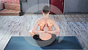 Fitness yoga man making namaste hands behind back sitting on mat in modern apartment