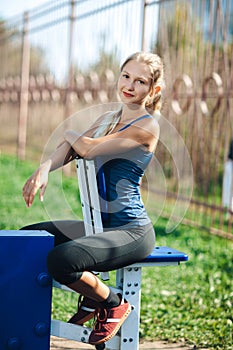 Fitness young woman in a blue shirt and leggings using outdoor gym equipment in the park looking at camera and smiling