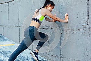 Rear view of fitness young woman doing stretching workout on a concrete grey wall. Athlete female exercising in morning outside