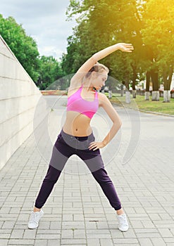 Fitness young woman doing warm-up stretching exercise before run, female athlete ready to workout in city park, sport and healthy