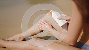 Fitted young woman applying waterproof sunscreen on legs at sandy beach. Pretty girl sits on sand with water resistant