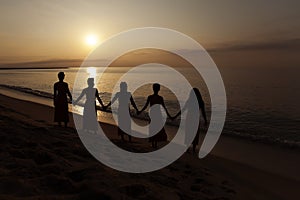 Five black women hold hands looking at the sunrise at the beach.