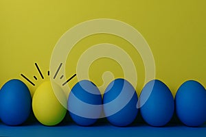 Five blue and a yellow funny faced painted Easter eggs stand on a yellow with blue background. Creative Easter concept