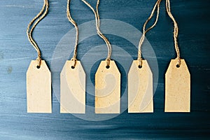 Five brown blank paper price tags or labels set hanging on a rope on the blue wooden background.