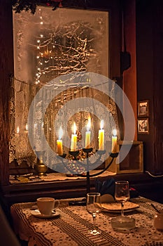 Five candles burning in menorah on a table on foreground and frosted window with snowed tree outside