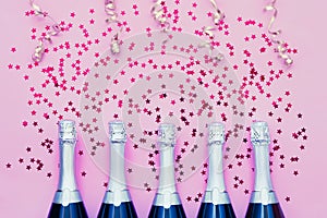 Five Champagne bottles with confetti stars and party streamers on pink background. Copy space, top view. Party background