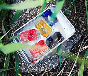 five cuvettes with watercolors in a tin box lie on a stone in the grass