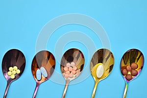 Five different types of pills in five multi colored metal spoons on a blue background with place for text