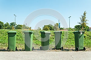 Five green plastic industrial garbage cans with junk and litter on the street in the city waiting for dumper truck to collect them