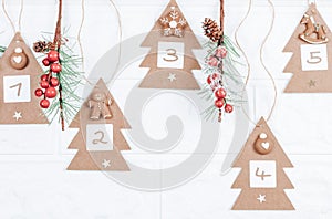 Five handicraft Christmas trees with numbers 1,2,3,4,5, cookies and spruce branches hang on a jute thread on a white.