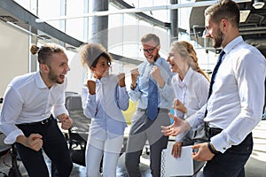 Five happy modern business people are keeping arms raised and expressing joyful while standing in large office