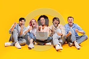 Five international students sitting with laptop and shaking fists in joy, reading great news over yellow background