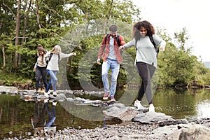 Five mixed race young adult friends hold hands and help each other while carefully crossing a stream standing on stones during a h